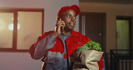 Pizza African American deliveryman in the red uniform and cap standing at the house with pizza boxes and calling on the phone in the evening. Outdoor.