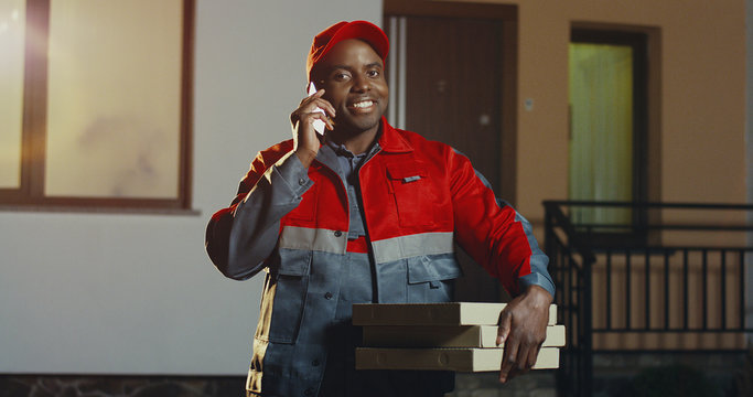 Pizza African American deliveryman in the red uniform and cap standing at the house with pizza boxes and calling on the phone in the evening. Outdoor. - Powered by Adobe