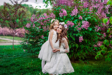 Obraz na płótnie Canvas Mom and daughter in wave hair hold hands near the lilac bush. On the head a rim of flowers. Dressed in white dresses.