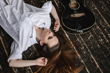 A brunette girl in a white shirt lies on a dark floor with a black guitar. Musician and vocalist searching for inspiration.