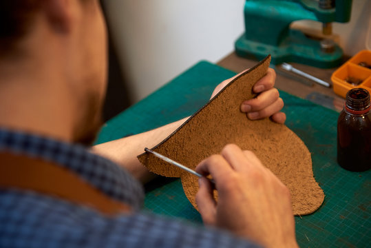 The master paints the edges of the leather pattern for the future product. Closeup of skinner craftsman working with brown natural leather using craft tools.
