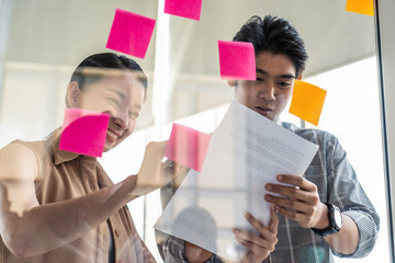 Asian young business people brainstorm to solve problem and planning new challenge topic. Woman show new idea on post it note at glass wall to man. Business finance and communication planning concept.