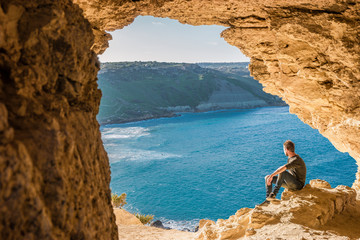 Gozo island Malta, young man in a cave looking out over the ocean and a View of Ramla Bay, from inside Tal Mixta Cave Gozo  - obrazy, fototapety, plakaty