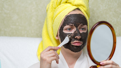 A girl with a towel on her head puts purifying black mask on her face by a cosmetic spatula looking in the mirror. Charcoal face mask or black clay mud. Skin care, acne treatment, cleansing skin.