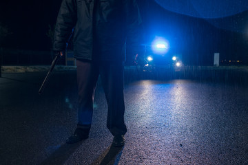 Police man with mask and nightstick and walkie talkie in hand watching and patrol car with sirens and blue lights on the curfew in the streets during the state of alarm in the covid 19 coronavirus.