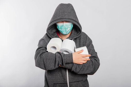 A young woman in a sweatshirt and a medical mask with a hood hides her face and holds rolls of toilet paper in her hands. Coronavirus panic concept, studio photo on white background.