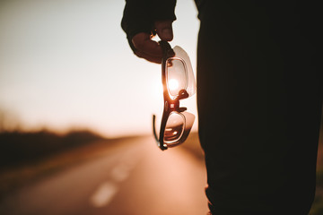 The biker stands on the road and holds his glasses in his hand