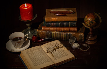 Naklejka na ściany i meble An old desk with a pile of vintage brown leather books, an open book with eye glasses, a cup of coffee, a red candle with flame, a vintage pocket clock, earth globe.