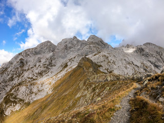 A pathway leading to high peaks in Italian Alps. Sharp slopes on both sides of the valley. Hard to reach mountain peaks. There are many mountain ranges in the back. Serenity and peace. Autumn vibes