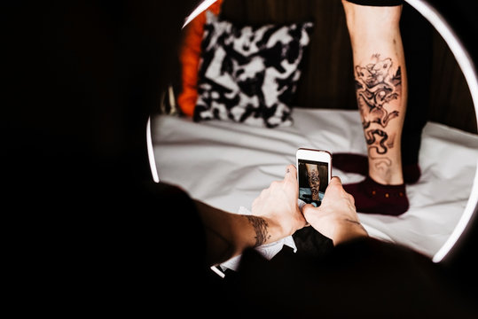 Unrecognizable man tattooer using smartphone to take picture of tattoo on leg of crop customer for portfolio in contemporary studio