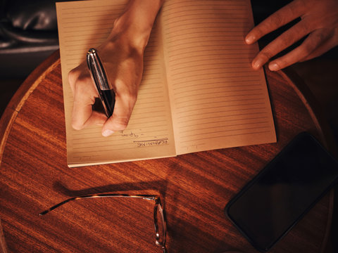 From above crop unrecognizable woman writing with pen in notebook at round wooden table with mobile phone and glasses in cafe