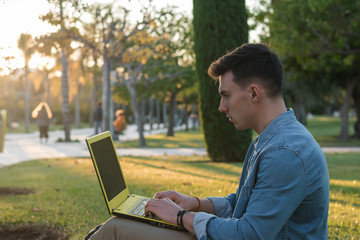 Side view of smart concentrated man with backpack studying at laptop sitting in park grass with crossed legs in sunny day