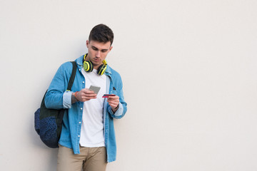 Smart stylish man in bright headphones surfing mobile phone and holding a credit card while leaning on marble wall in sunny day