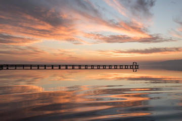 Silhouettes of distant unrecognizable people standing on pier near tranquil lake against sunset sky