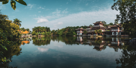 View of calm lake water reflecting sky and oriental buildings on shore with lush tropical greenery, Qingxiu Mountain, China