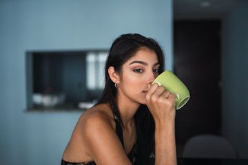 Beautiful passionate brunette Hispanic woman in dress looking at camera and holding mug of hot drink at table with volume in room