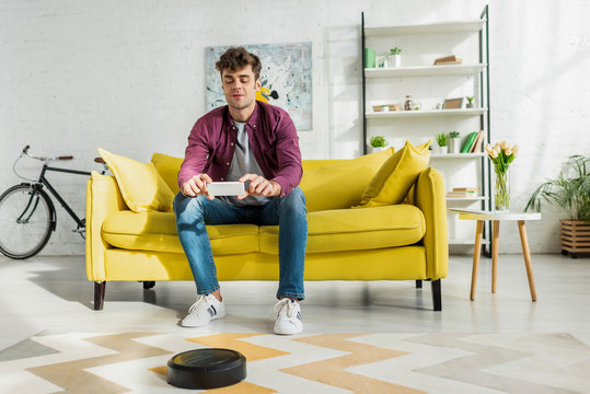 cheerful man taking photo on smartphone while robotic vacuum cleaner washing carpet in living room