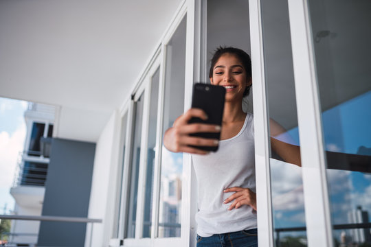 Beautiful brunette Hispanic woman taking selfie on mobile phone and standing between balcony windows