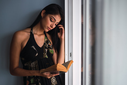 Beautiful passionate brunette Hispanic woman in dress looking at volume in hands near window and grey wall