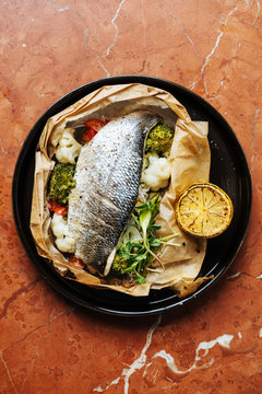 Top view of baked in paper fish with broccoli cauliflower herbs and piece of lemon on table in restaurant 