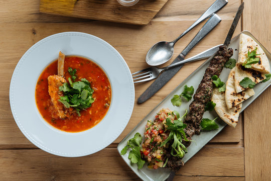 Top view of red soup with meat and fresh herbs on wooden table with kebab and flat bread in restaurant