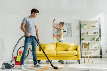 happy man using vacuum cleaner near woman waving hand in living room