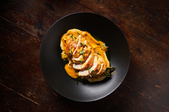 Top view of baked sliced chicken fillet with colorful orange sauce and green herbs in stylish black bowl on wooden table 