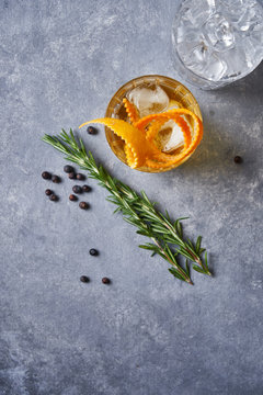From above top view of glass cup with cold old fashioned cocktail with whiskey and orange peel placed on grey table with rosemary plant a pepper grains