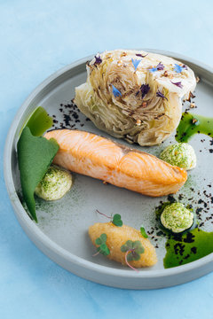 From above of baked salmon fillet and pike caviar with piece of young cabbage on stylish grey plate decorated with white sauce on blue background