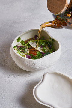 Unrecognizable person pouring fresh warm tea from glass teapot into ceramic bowl with herbs