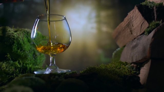 slow motion. pours  cognac or brandy into a glass against the backdrop of nature trees and sunset. firewood moss nature. wood. evening drinking in nature. close up