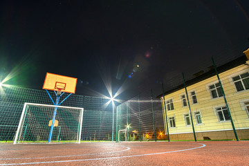 Outdoors mini football and basketball court with ball gate and basket surrounded with high protective fence brightly illuminated with spotlight lamps at night. © bilanol