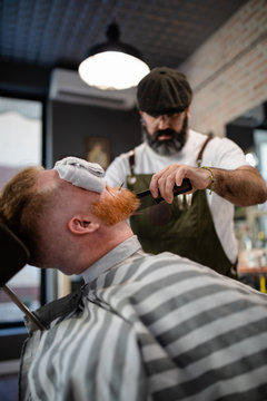 Blurred unrecognizable barber with comb fixing redhead man beard with towel covering eyes sitting in barbershop