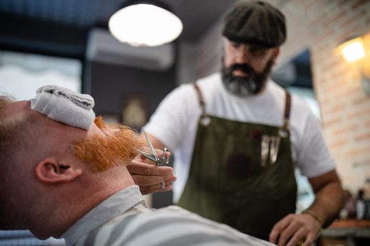 Blurred unrecognizable barber with scissor fixing redhead man beard with towel covering eyes sitting in barbershop