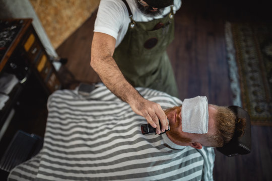 From above top view of cropped unrecognizable barber with trimmer cutting redhead man beard with towel covering eyes sitting in barbershop