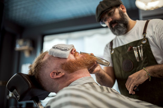 Barber with comb and trimmer cutting redhead man beard with towel covering eyes sitting in barbershop