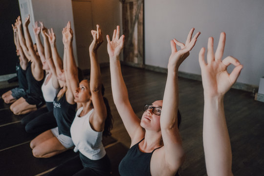 High angle of group of young sporty women and men in sportswear with arms raised and Gyan Mudra stretching body and meditating while sitting in virasana pose on sports mats and looking up training in modern yoga studio