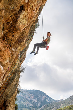From below athletic female alpinist hanging on cliff under cloudy sky