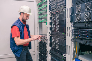 An engineer in a white helmet connects Internet wires to new equipment. A man works in the server room of a data center. A technician connects a cable to a stack of managed switches. 