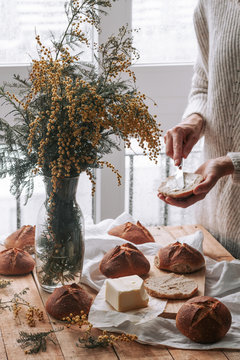From above of crop woman spreading butted on homemade bread with knife on table with vase of yellow field flowers