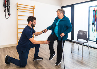Young bearded male personal trainer kneeling while holding hand and knee of aged woman during exercising with cane in gym 