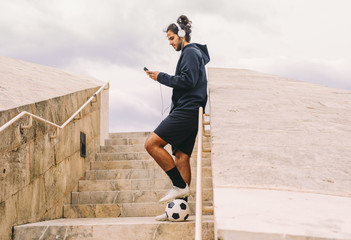 From below side view of modern male soccer player listening to music with headphones and smartphone while standing with ball on stairs in city in summer day with grey cloudy sky in background