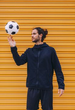 Young bearded male soccer in dark blue sports hoodie and shorts spinning ball on finger while standing against yellow roller shuttle background