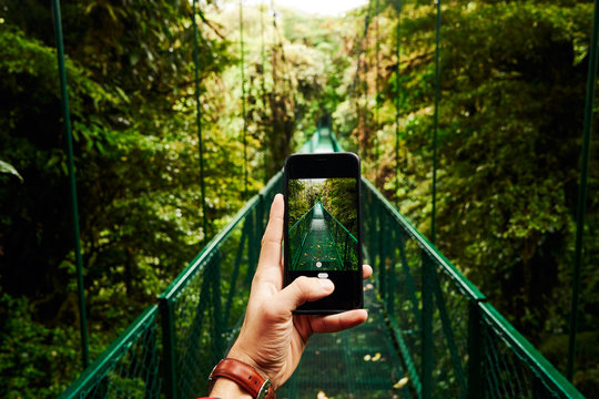 Unrecognizable traveler using smartphone to take picture of bridge going through green jungle during journey in Costa Rica