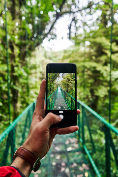 Unrecognizable traveler using smartphone to take picture of bridge going through green jungle during journey in Costa Rica