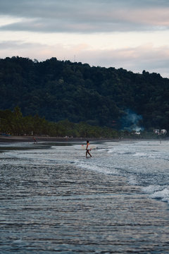 Unrecognizable man with surfboard walking near waving sea in cloudy evening on beach in Costa Rica