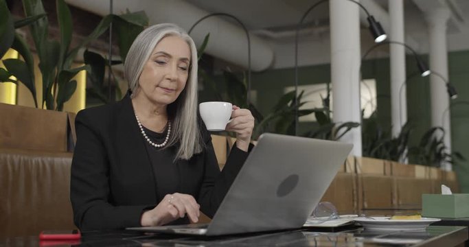 Front view of mature stylish businesswoman holding cofee cup and working on laptop. Old elegant female enterpreneur sitting at cafee table and working. Indoor. Concept of business.