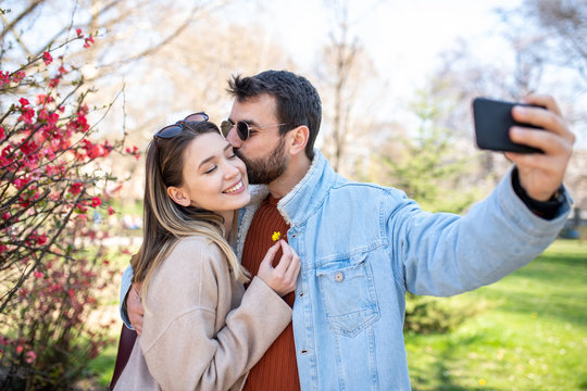 Man and woman taking a cute selfie outside, while the man is kissing the womans cheek.
