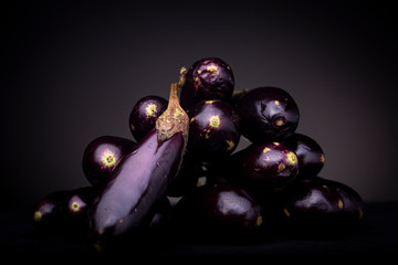 Small vibrant purple baby eggplant leaning on a pile of dark aubergine vegetable. Studio low key food image on black surface and dark grey background.