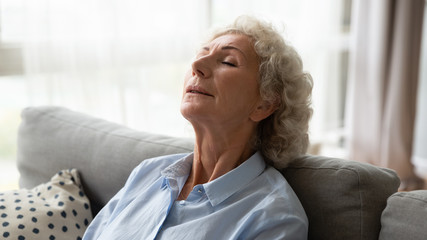 Relaxed elderly woman sit rest on cozy couch in living room take nap daydream at home, calm mature female grandmother sleep relax on comfortable sofa breathing fresh air, stress free concept